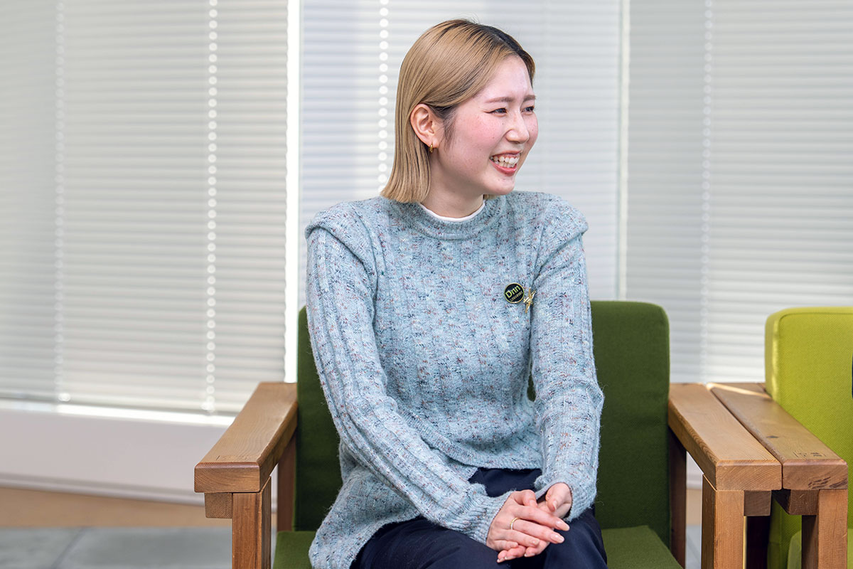 ブラインドを背景に椅子へ座る藤目の写真。両手を重ねて笑顔を見せ、金髪ボブヘアと小さなピアスが印象的である。落ち着いた室内で談笑している様子である