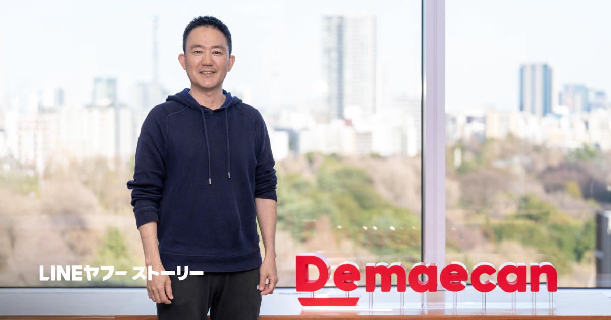 大きな窓の前に立つ出前館CEO矢野さんの全身写真。ネイビーのパーカーを着て微笑み、背後には都市の景色と「Demaecan」の赤いロゴが見える。左下に白文字「LINEヤフーストーリー」がある。