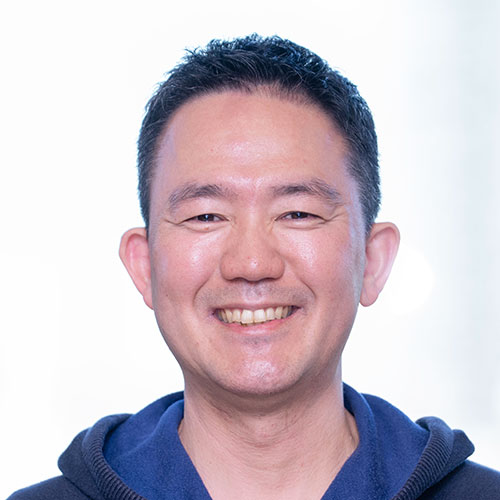 男性の顔のクローズアップ。短髪で明るい笑顔を見せ、ネイビーのパーカーを着て正面を見つめている。