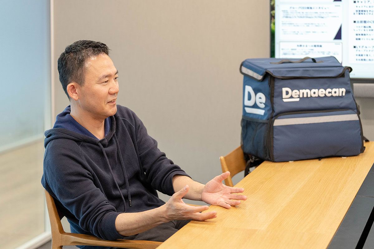 テーブルで身振りを交えて話す男性の写真。ネイビーのパーカー姿で両手を広げ、隣に配達用バッグが置かれている。