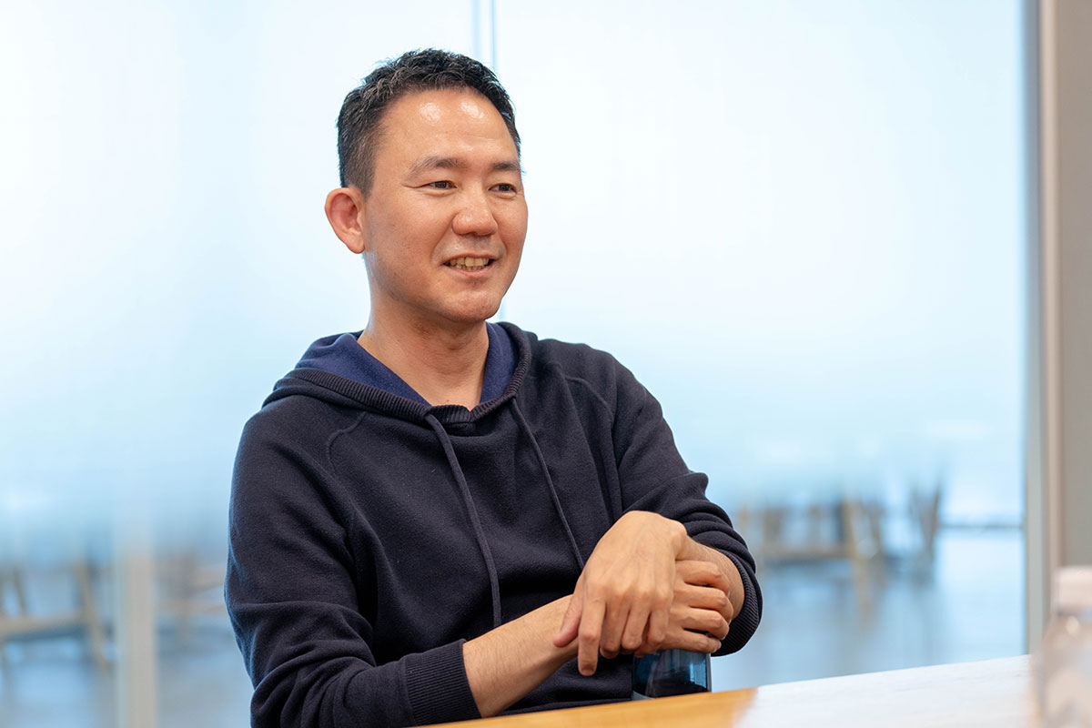 テーブルに座る出前館CEOの上半身写真。ネイビーのパーカー姿で腕を軽く組み、穏やかな表情で会話している。背景には窓と屋外の景色が広がる。