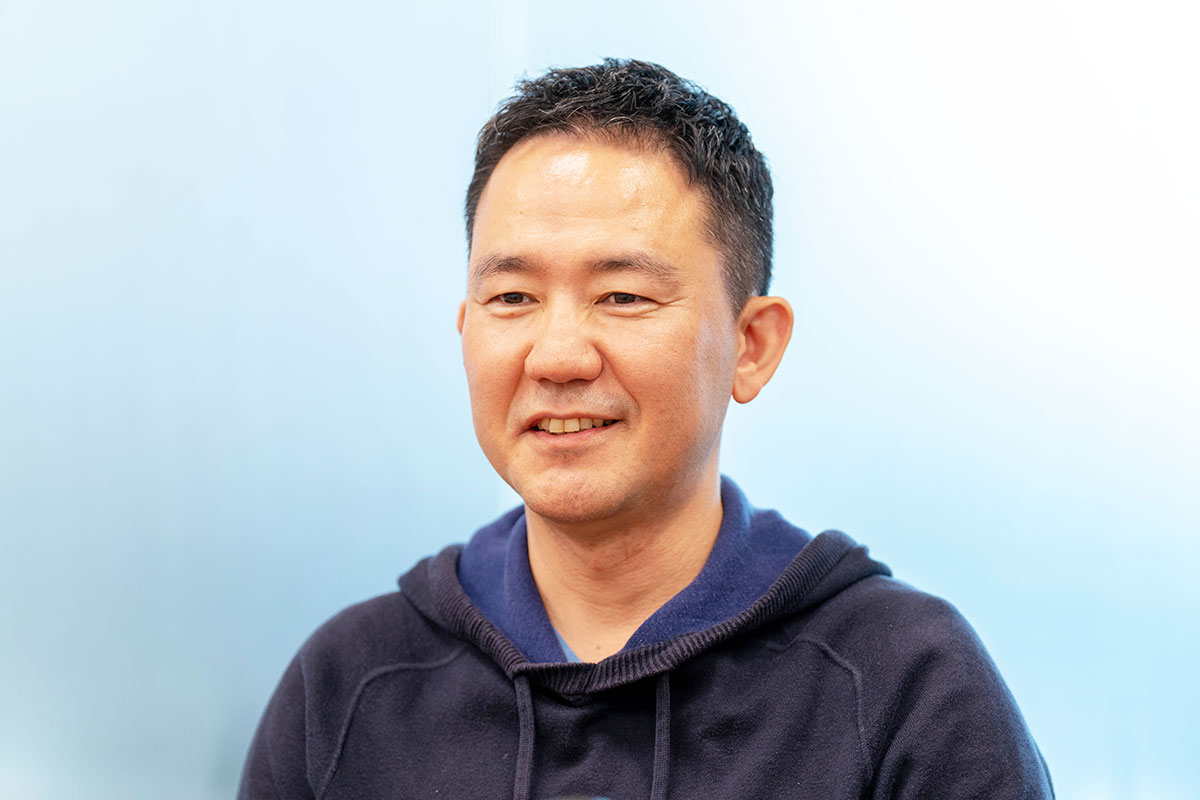 出前館CEOのバストショット。ネイビーのパーカーを着て柔らかく微笑み、明るい室内でリラックスした雰囲気を見せている。