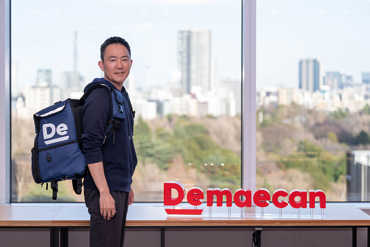 大きな窓の前でリュックを背負った出前館CEOの写真。ネイビーのパーカー姿で振り返り、赤い「Demaecan」ロゴの横に立っている