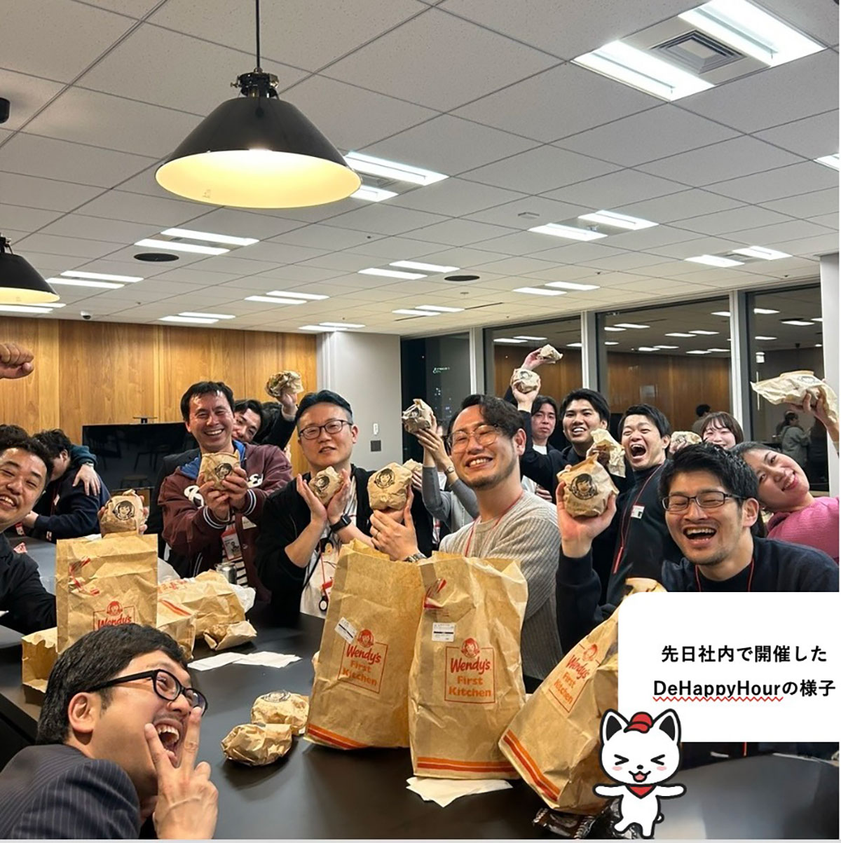 会議室で大勢が集まり食事を楽しむ写真。紙袋やハンバーガーを手に笑顔を見せる人々が並び、賑やかな雰囲気で交流している様子である。