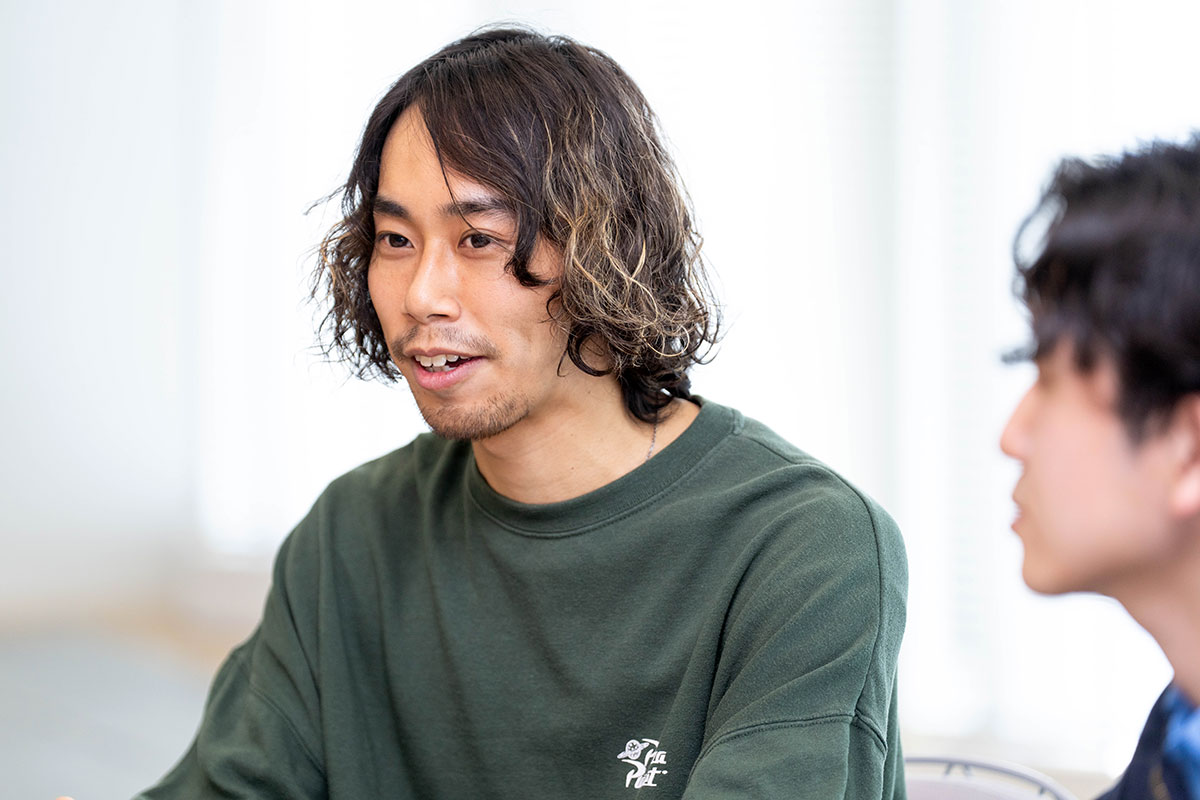 緑のトップスを着た男性、百瀬の上半身写真。ゆるいパーマの髪とあごひげが特徴で、前方を見ながら口を開き、真剣に話している様子である。