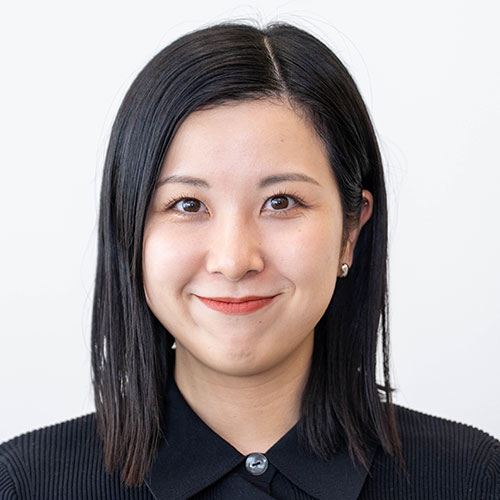 グレー背景の前で微笑む女性、山口のバストショット。黒髪のストレートボブに黒のトップスを合わせ、小さなピアスを着け、口元に笑みを浮かべている。