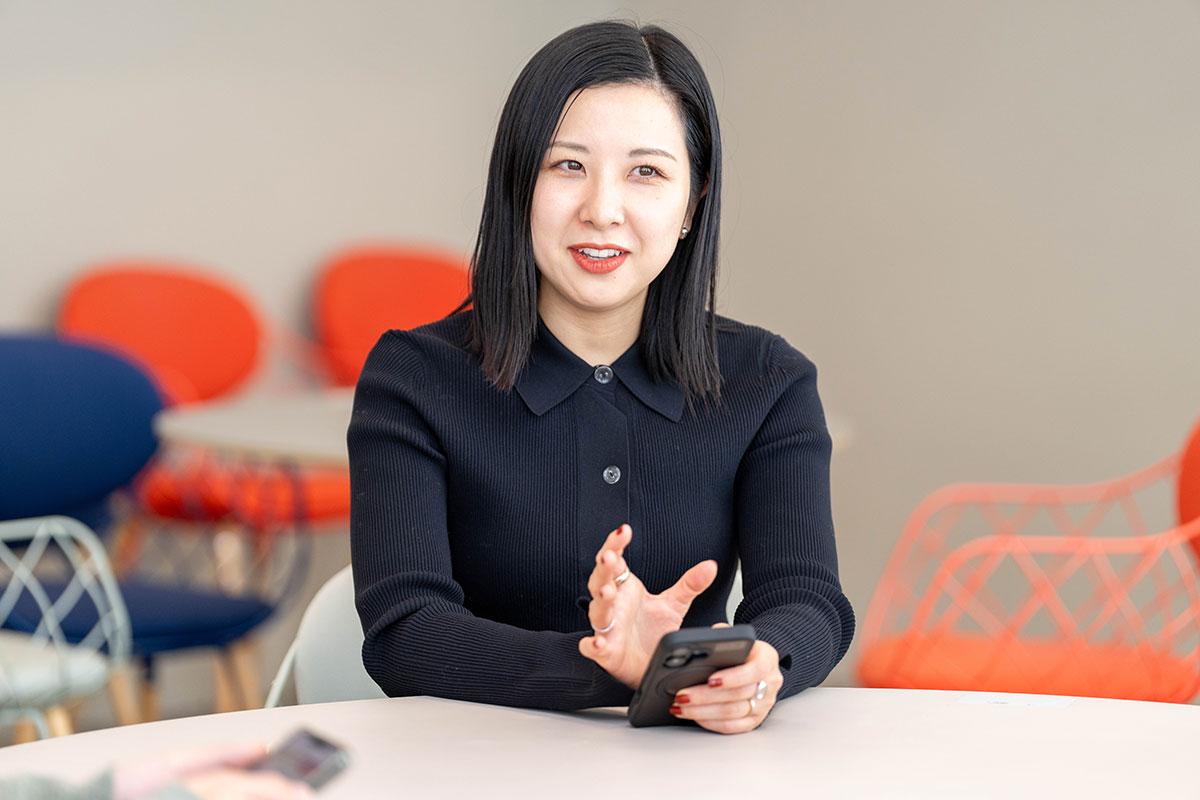 室内で話す女性、山口の上半身写真。黒の長袖トップスを着てテーブルに座り、少し首を傾けながら真剣な表情で会話している。背後に赤や青の椅子がぼけて見える。