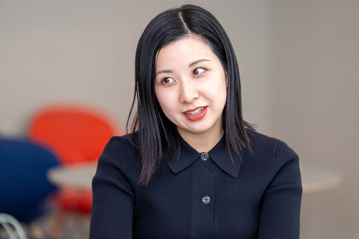 テーブルに座る女性、山口がスマートフォンを手にし、片手を上げながら説明する写真。黒のトップス姿で落ち着いた表情を見せ、明るい室内で会話している様子である。