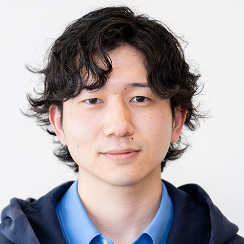 グレー背景の前で正面を向く男性、山口のバストショット。黒のウェーブがかった髪を分け、青いシャツに濃紺のパーカーを重ね、落ち着いた表情でカメラを見ている。