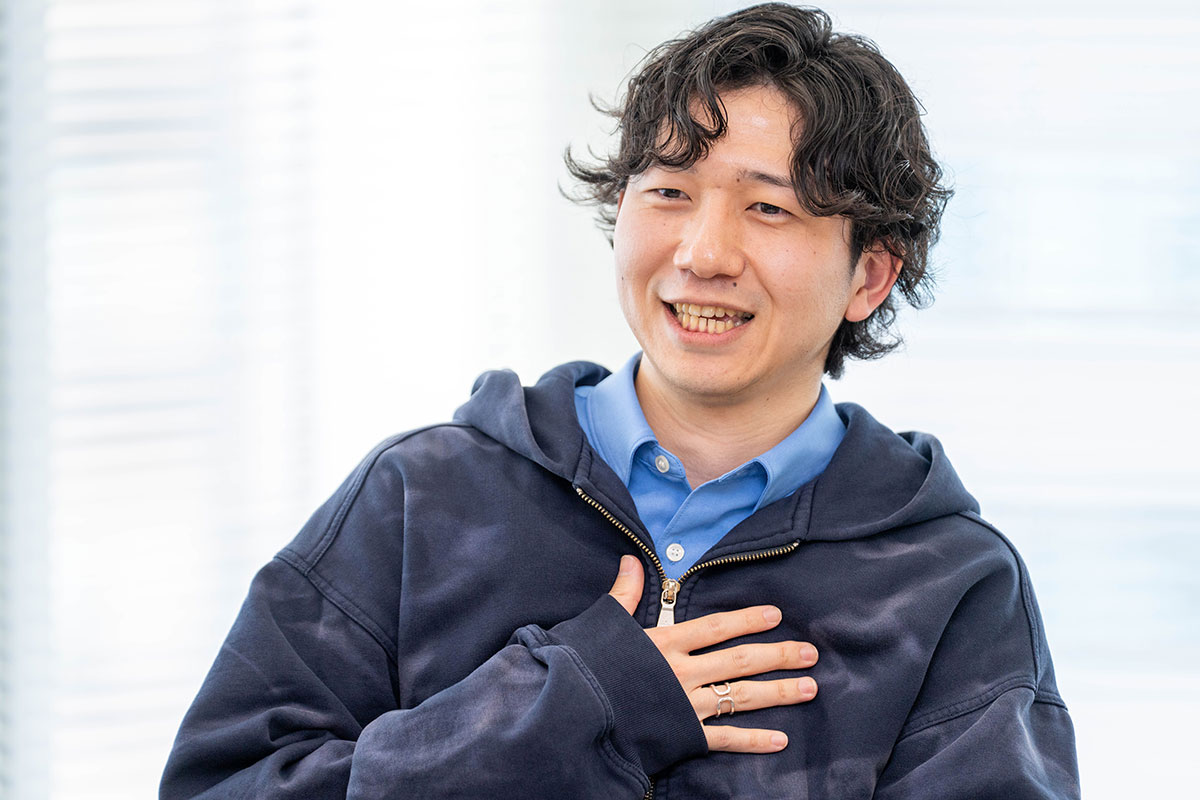 青いシャツに紺のパーカーを重ねた男性、山口の写真。胸に手を当てて笑顔を見せ、和やかな雰囲気で会話している。背景は白いブラインドである。