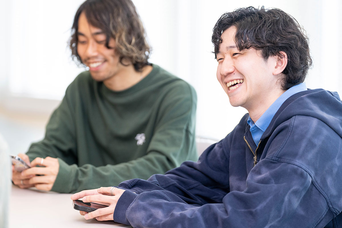 テーブルに並んで座る男性二人の写真。手前の男性は青いシャツと紺のパーカー姿で笑い、奥の男性は緑のトップスでスマートフォンを持ちながら微笑んでいる。