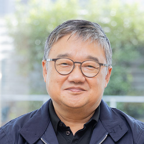 A bust shot of CEO Fan facing the camera with a calm expression. He has short gray hair, wears round glasses, and is dressed in a dark navy jacket. Soft greenery is blurred in the background, with gentle natural light falling on him.