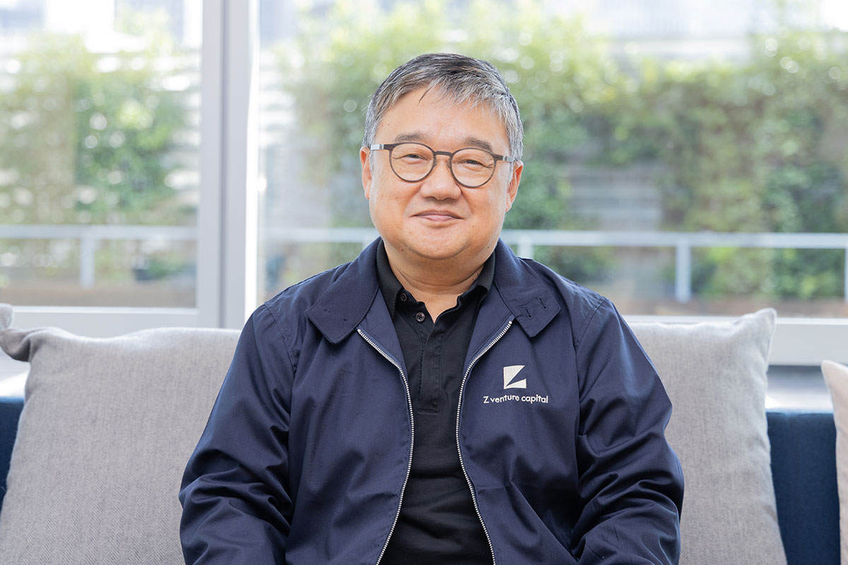 A front-facing photo of CEO Fan seated on a sofa. He is wearing a navy jacket and has a composed expression. Greenery spreads out beyond the large window behind him, creating a calm and gentle atmosphere in the bright interior.