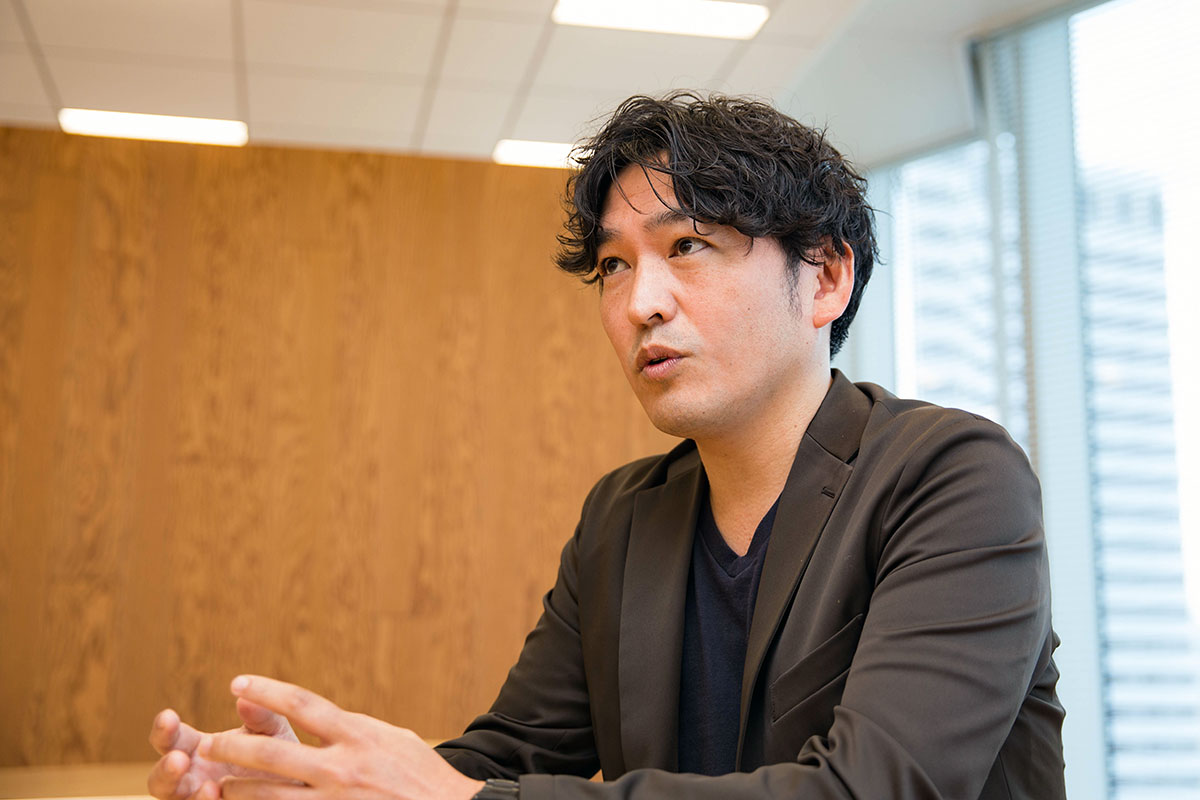 男性、小用が木目の壁を背景に座り、両手を前に出して語っている。黒いジャケットを着て真剣な表情をしている。室内は明るく、横には大きな窓があり、外のビルが少し見えている。