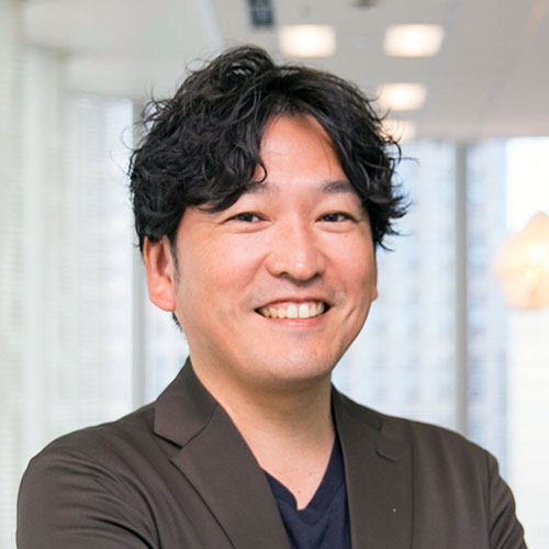 男性のバストショットで、黒髪のウェーブヘアが特徴的だ。黒いインナーに濃いジャケットを着て笑顔を見せている。明るいオフィスの窓辺で撮られ、背景にはビル群がぼんやり映っている