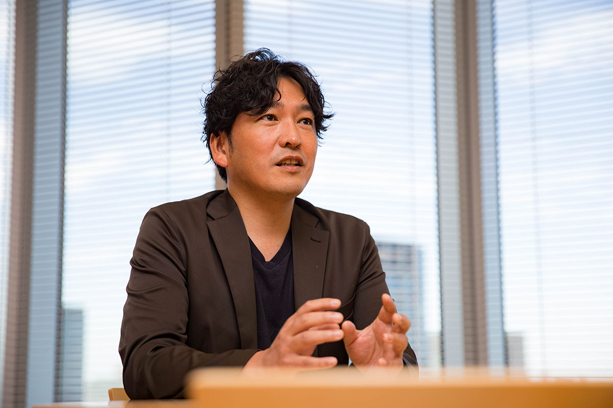 男性がテーブル越しに話している様子で、手を前に出し説明しているように見える。黒髪のウェーブヘアで黒いジャケットを着ている。背景の大きな窓にはブラインドがかかり、青空とビルがぼんやり映っている。