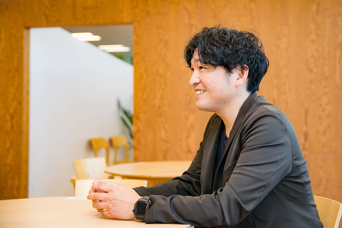 男性が明るい木目調の室内で微笑みながら座っている。黒髪のウェーブヘアで黒いジャケットを着ており、両手をテーブルの上で軽く組んでいる。奥には椅子や観葉植物が見え、落ち着いた空間となっている。