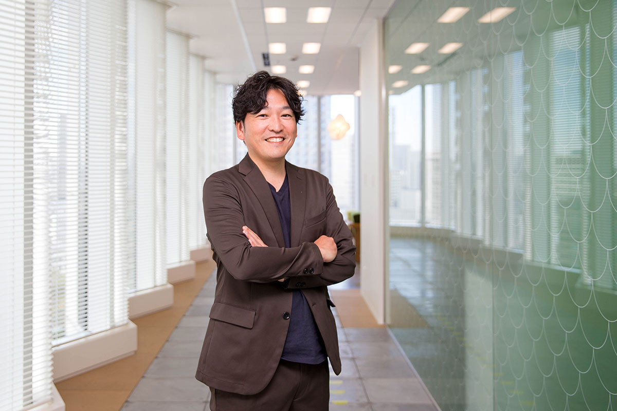 オフィスの廊下で腕を組んで立つ男性が笑顔を向けている。黒いインナーにダークブラウンのジャケットを着ている。左側の窓から光が入り、右側のガラス壁には模様が映り込んでいる。