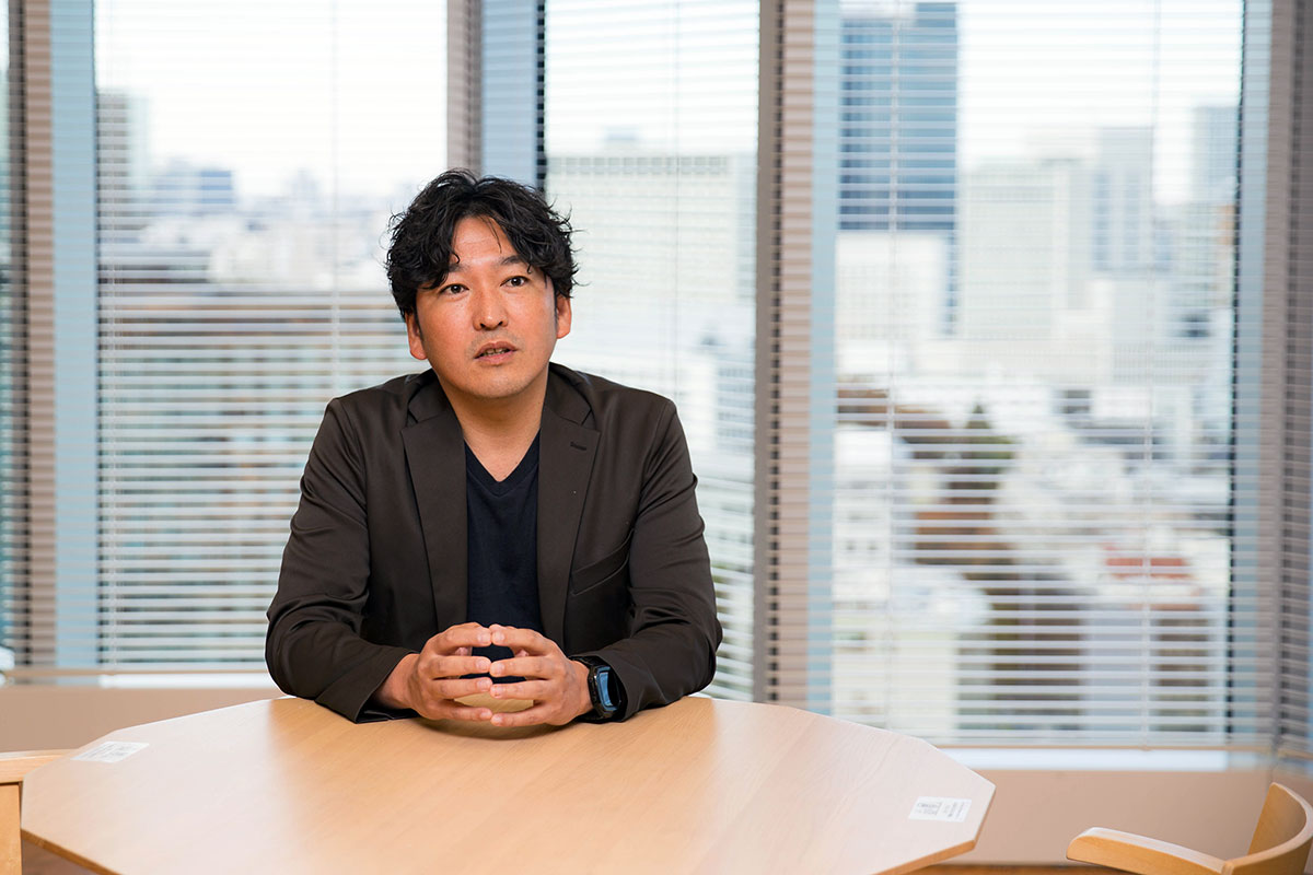 小用が円形のテーブルに座り、手を組んで前方を見る。黒いジャケットとTシャツを着て落ち着いた表情だ。背景の大きな窓の向こうに都市のビル群がぼんやりと広がっている。