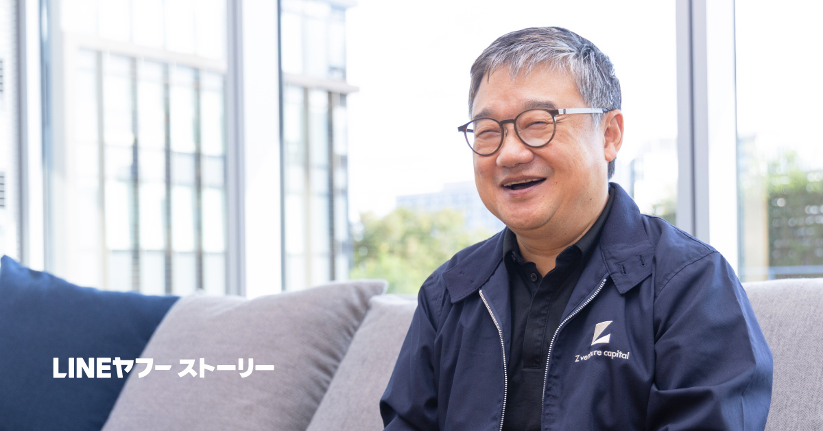 ソファに座った男性、ファンCEOが笑顔でこちらを向いている。短いグレーの髪と丸い眼鏡をかけ、紺色のジャケットを羽織っている。背景の大きな窓越しにビルが見え、画面左下に白字で「LINEヤフー ストーリー」と表示されている。