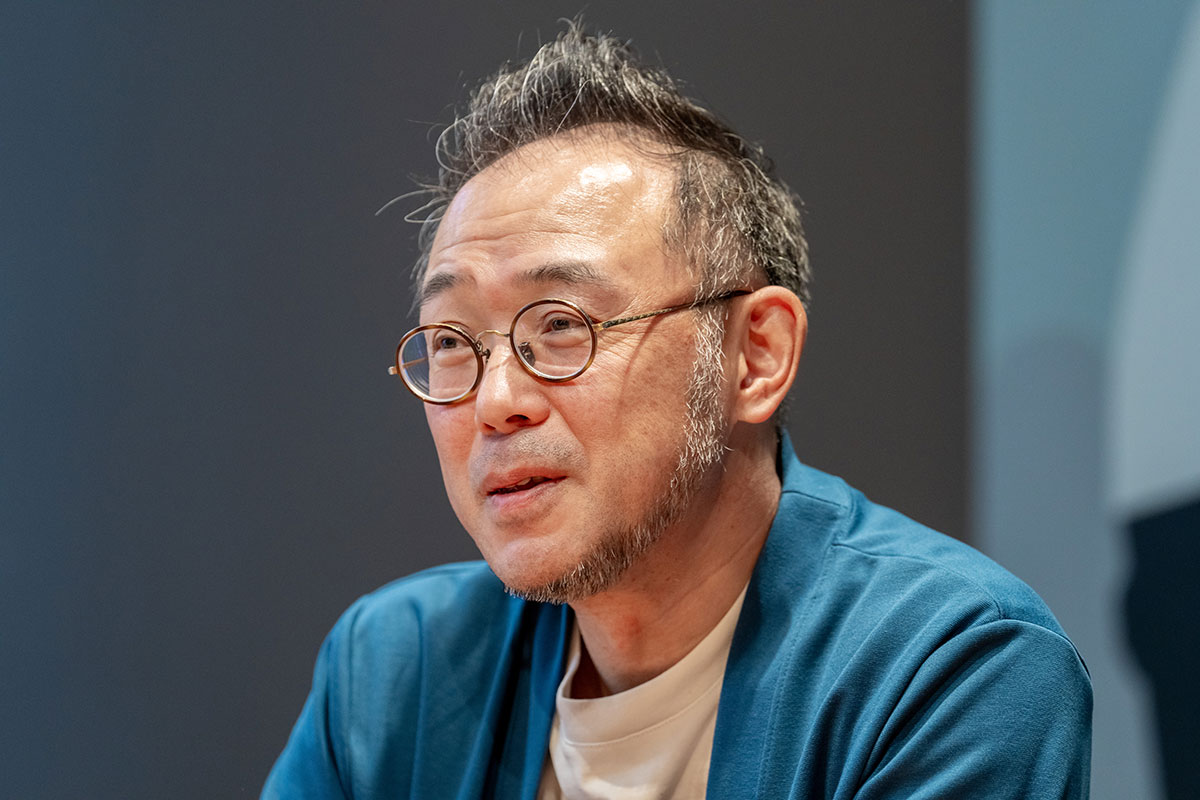 丸眼鏡の澤田CEOが斜めを向いて話している様子。青い羽織風の上着に白いTシャツを合わせ、落ち着いた表情を浮かべている。背景は暗めの室内