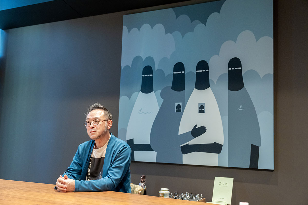 会議室風の部屋で丸眼鏡の澤田CEOが机に座っている。後ろには雲を背景に黒いフードを被った人物を描いた大きなイラストが飾られている