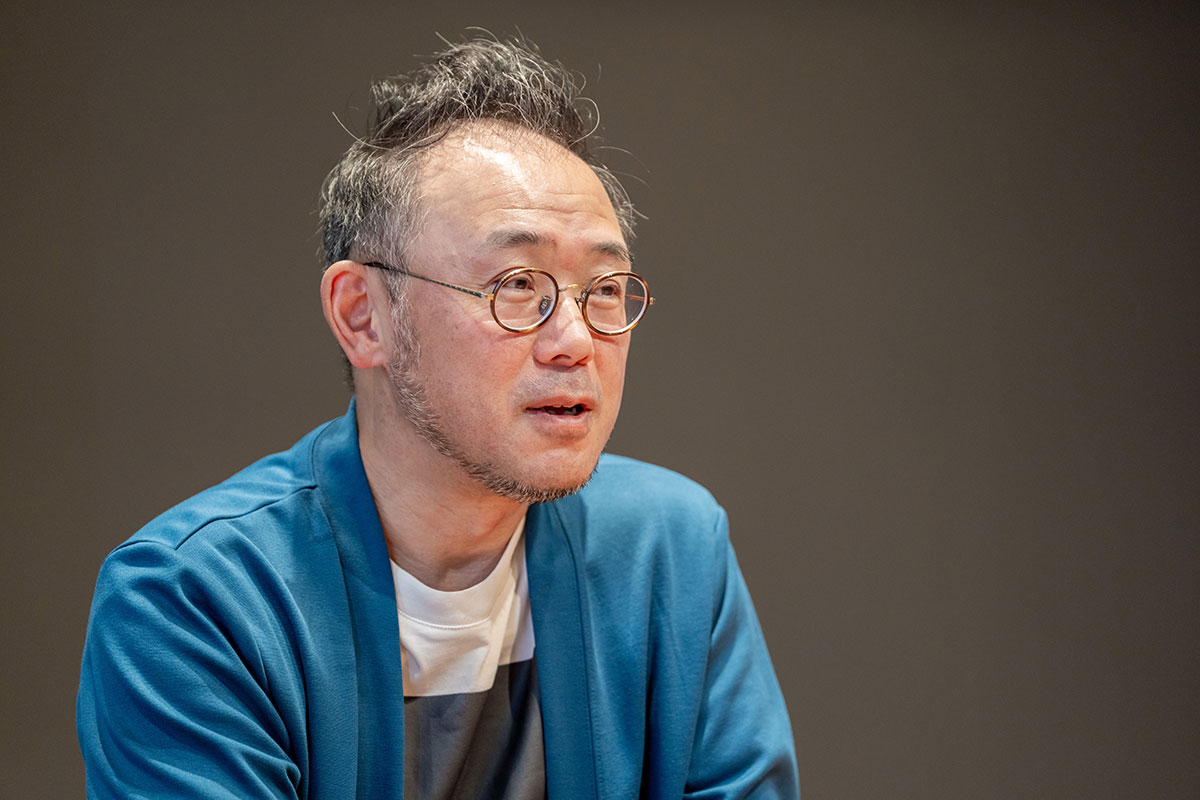 会議室風の部屋で丸眼鏡の澤田CEOが机に座っている。後ろには雲を背景に黒いフードを被った人物を描いた大きなイラストが飾られている