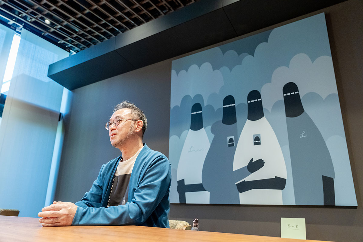 青い羽織を着た澤田CEOが会議机に座っている。背景には黒いフードの人物を描いた大きなイラストが壁に掛けられており、現代的な空間の雰囲気