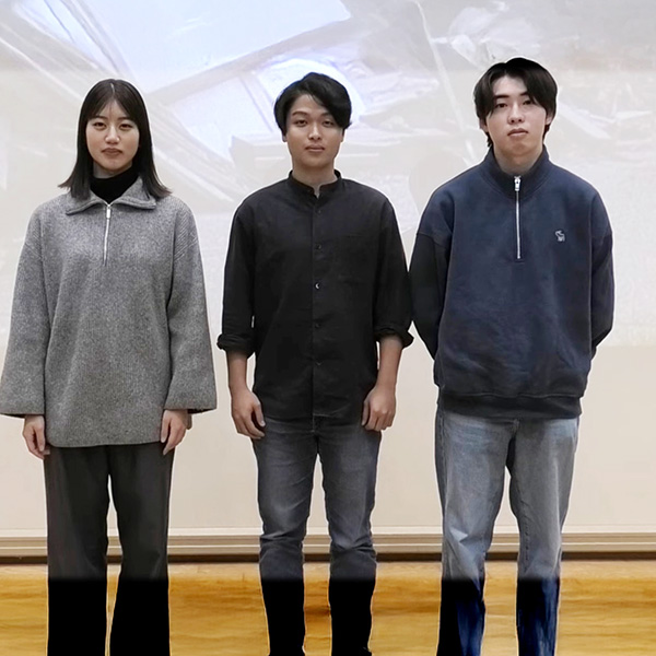 三人の大学生が並んで立っており、背景には何かの映像が映し出されています。