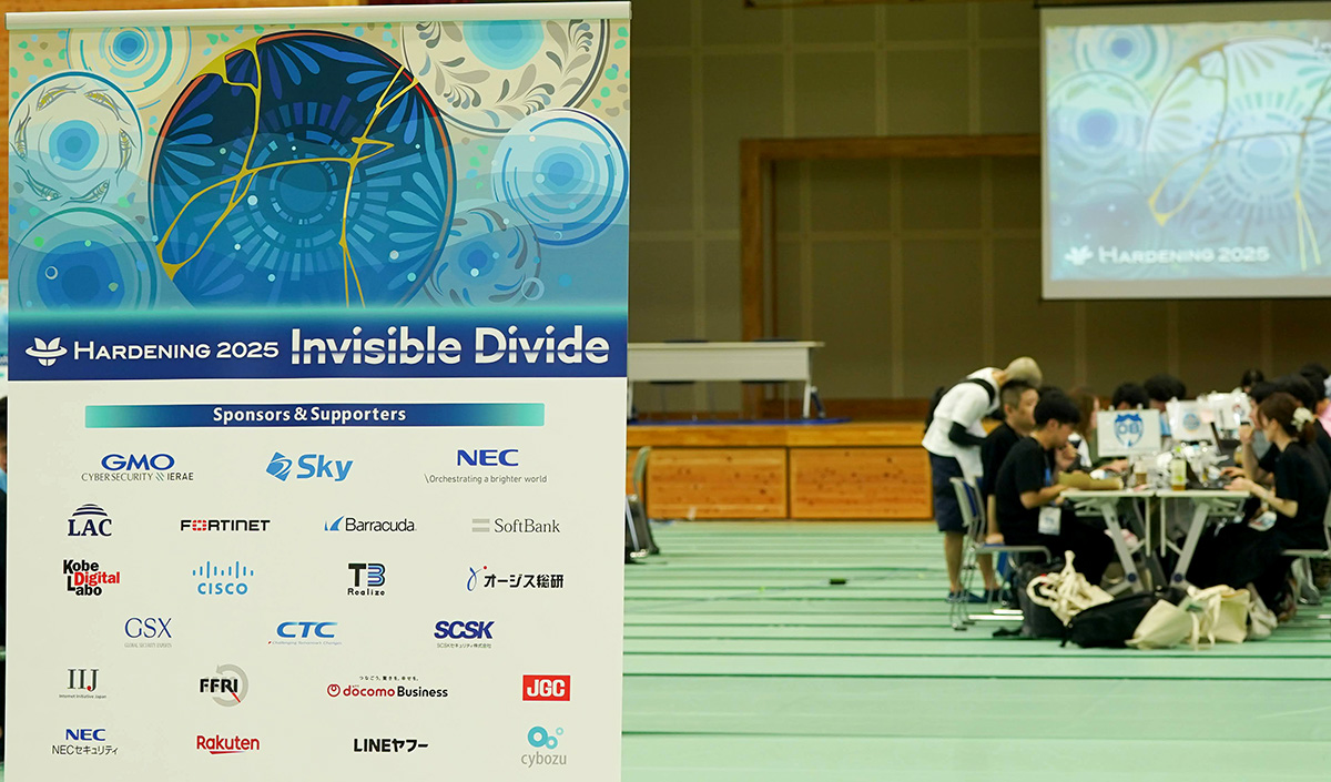 体育館のような広い室内で、イベント「HARDENING 2025 Invisible Divide」の立て看板が大きく写っている。看板には青を基調とした抽象的なデザインが描かれ、下部には「GMO」「Sky」「NEC」「SoftBank」「楽天」「LINEヤフー」など、多数のスポンサー企業のロゴが並んでいる。