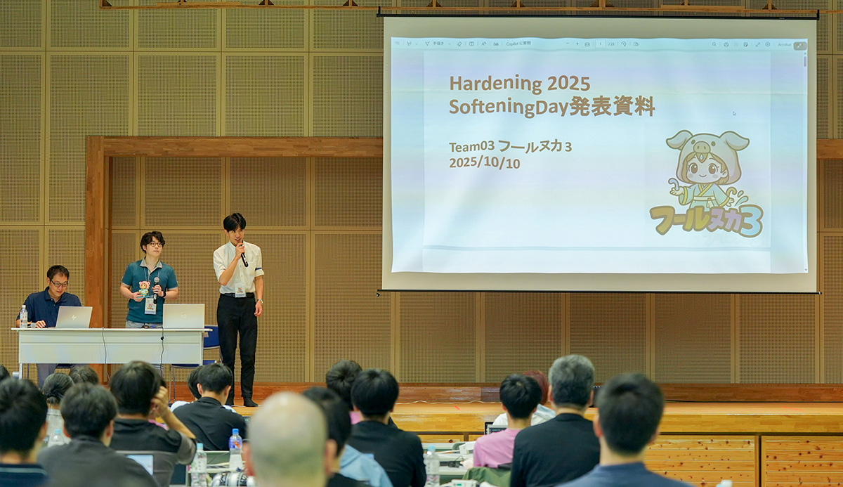 室内のステージ上で、3人の発表者が登壇してプレゼンテーションを行っている様子。中央のスクリーンには「Hardening 2025 Softening Day 発表資料」と表示され、右側に着ぐるみ風のキャラクターが描かれている。