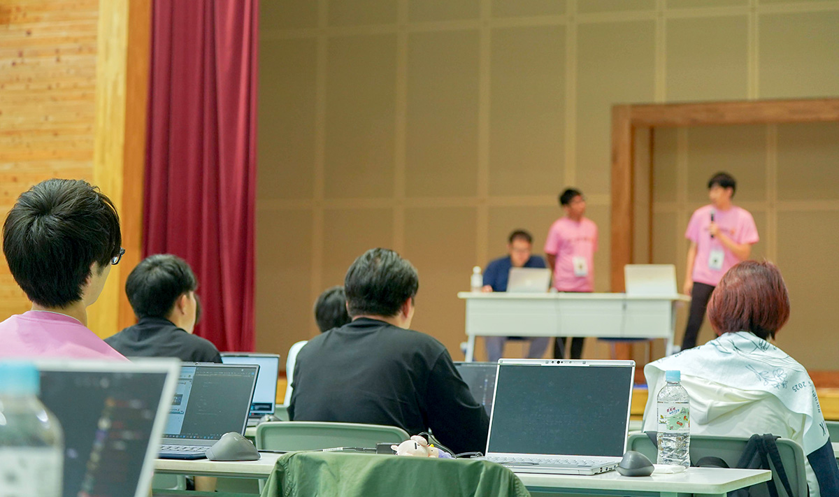 室内の会場で、観客席からステージ上の発表者たちを見ている様子。前方のステージにはピンクのTシャツを着た2人の発表者が立ち、マイクを使って説明している。