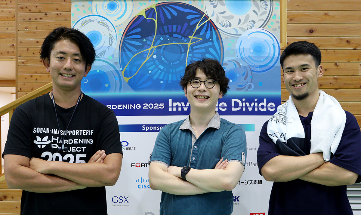 「HARDENING 2025 Invisible Divide」と書かれたイベントバナーの前で、3人の男性が並んでポーズを取っている様子。全員が腕を組み、笑顔でカメラを見ている。