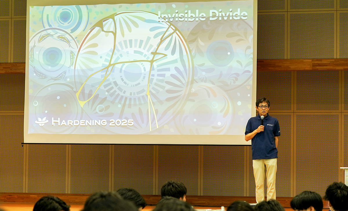 室内のステージ上で、マイクを手にした男性が登壇して話している様子。背後のスクリーンには「HARDENING 2025 Invisible Divide」と書かれ、円や線が重なった抽象的な青系のデザインが投影されている。