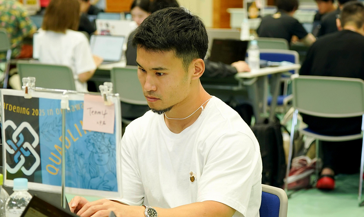 室内の会場で、白いTシャツを着た男性がノートパソコンを操作している様子。短髪で整えられた髪型をしており、真剣な表情で画面を見つめている。