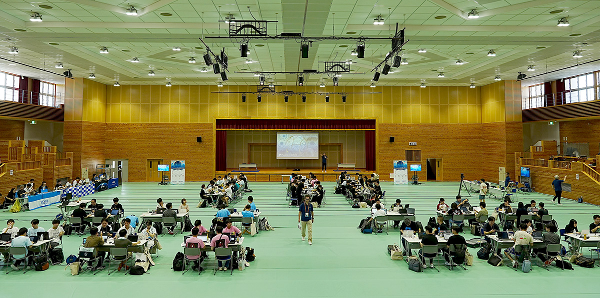 体育館のような広い会場で、多くの人々が円卓や長机に分かれて着席し、ノートパソコンを使いながら作業している様子。中央奥のステージ上にはスクリーンが設置され、「HARDENING 2025 Invisible Divide」のロゴが映し出されている。