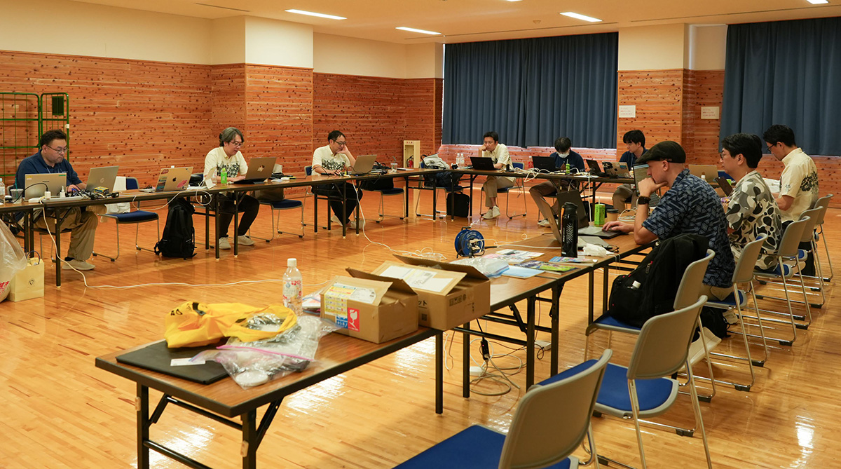 目調の壁と床の会議室で、十数名の人々が円形に並べられた長机に座り、ノートパソコンを操作している様子。多くの人がTシャツ姿で、イヤホンをつけたり、画面に集中したりしている。