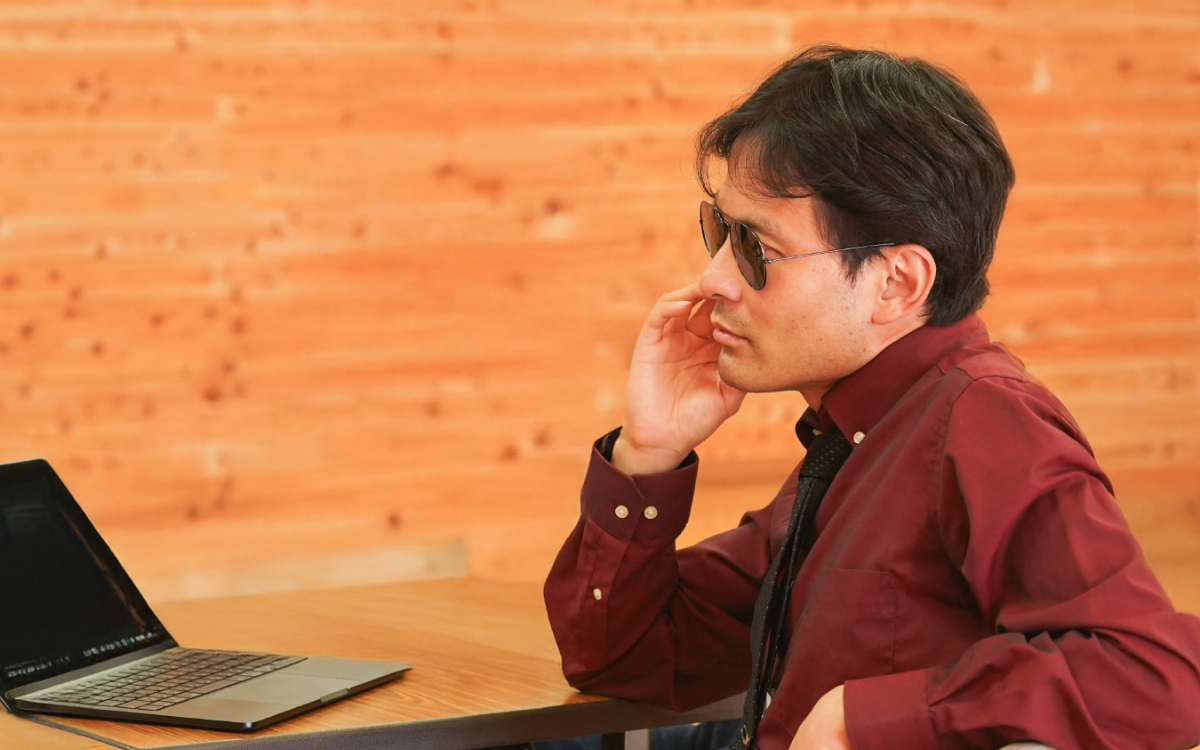 木目の壁を背景に、ボルドー色のシャツと黒いネクタイを着た男性が座っている。サングラスをかけ、頬に手を添えて考え込むような姿勢をしている。
