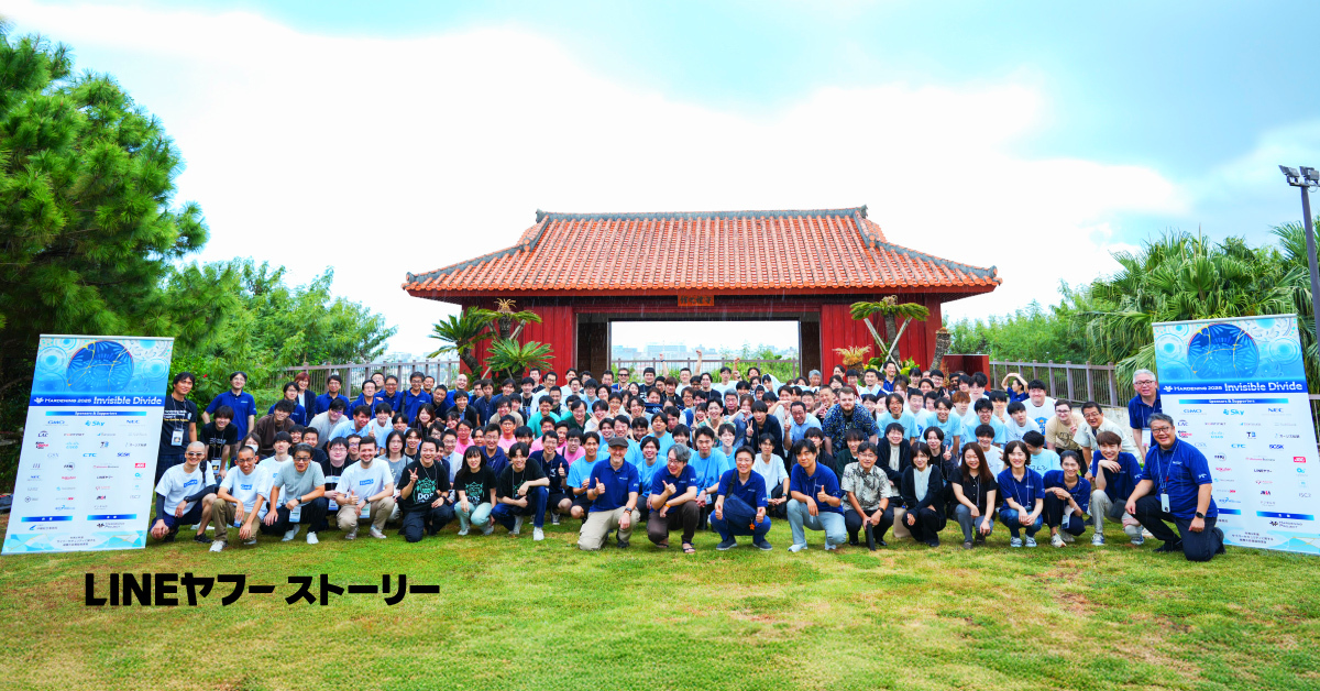 赤瓦の伝統的な門を背景に、大勢の人々が屋外で集合写真を撮っている様子。人々は前列がしゃがみ、後列が立っており、全体で100人以上が並んでいる。左右にはイベントスポンサーのロゴが並ぶ立て看板が設置されており、記念撮影の場であることがわかる。