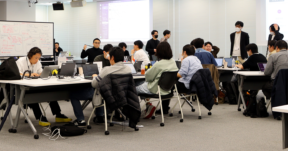 会議室で複数の人がノートパソコンを使いながらグループ作業をしている様子の写真。長テーブルを囲んで多くの参加者が座り、パソコンを操作したり、周囲の人と会話したりしている。