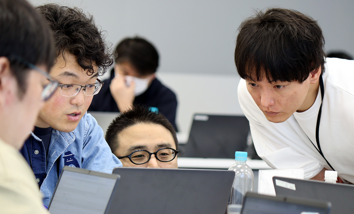 会議室で複数の男性がノートパソコンの画面を見ながら議論している様子。