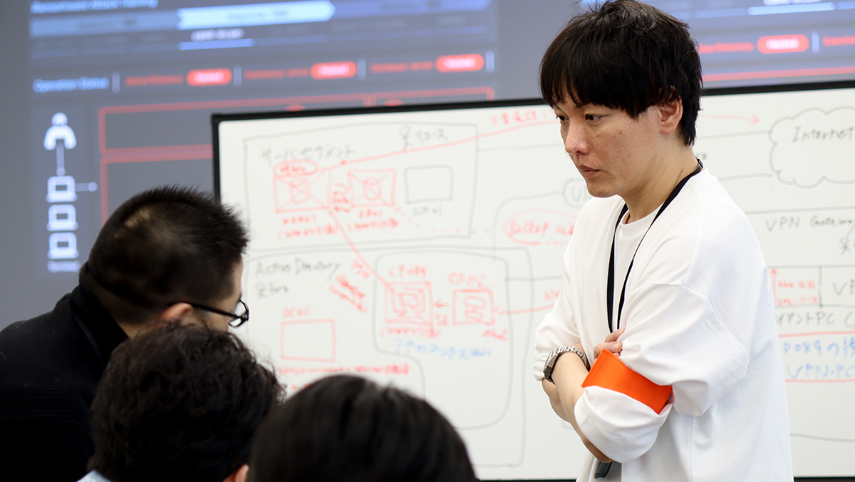 会議室で男性が腕を組みながら、前にいる参加者たちの議論を真剣な表情で見つめている様子。