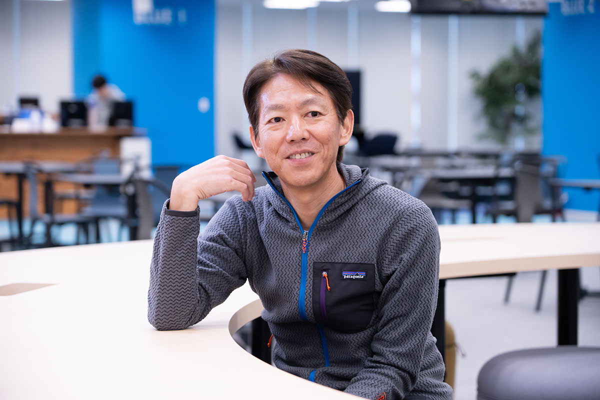 オフィスのような場所で、榊社長がテーブルに肘をついてリラックスして座っています。
