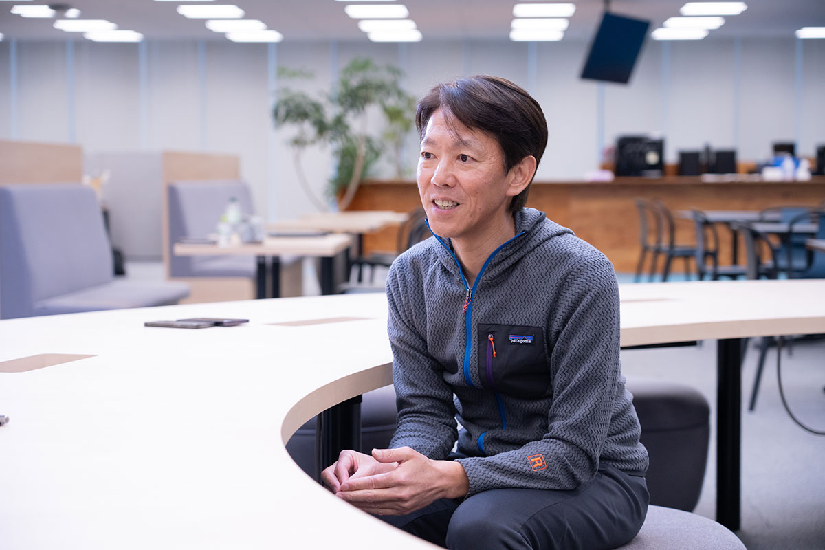 榊社長がテーブルに座り、話をしているような姿勢です。背景にはオフィスの家具があります。