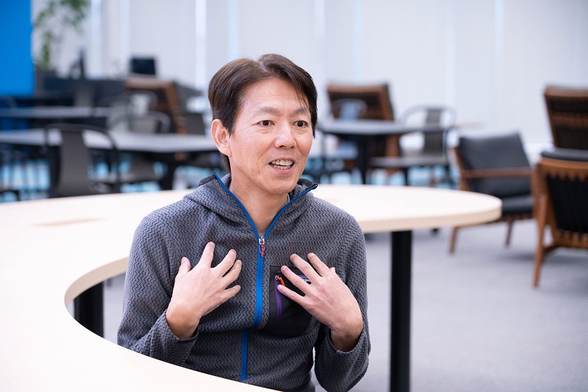 榊社長が両手を胸に置き、話をしている場面です。背景にはオフィスのインテリアが見えます。