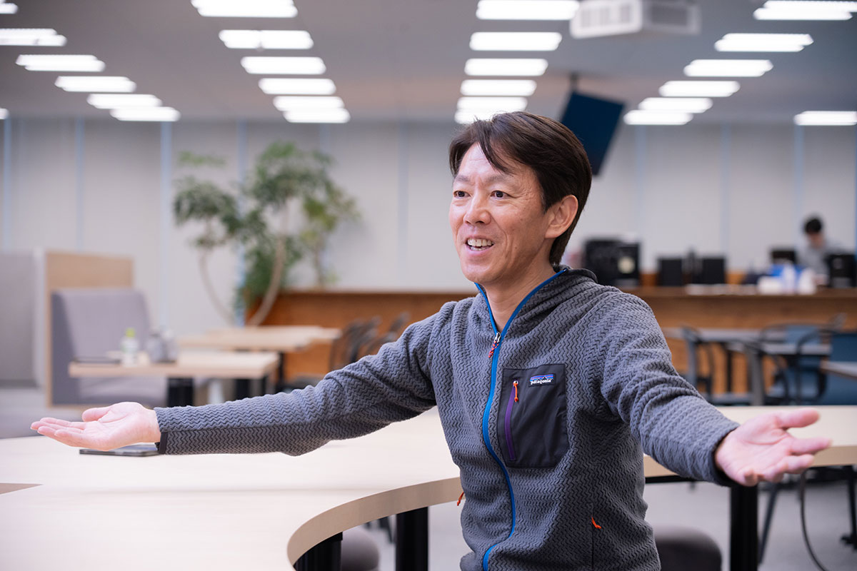 榊社長が両手を広げて笑顔で話している姿です。背景にはオフィスの様子が見えます。