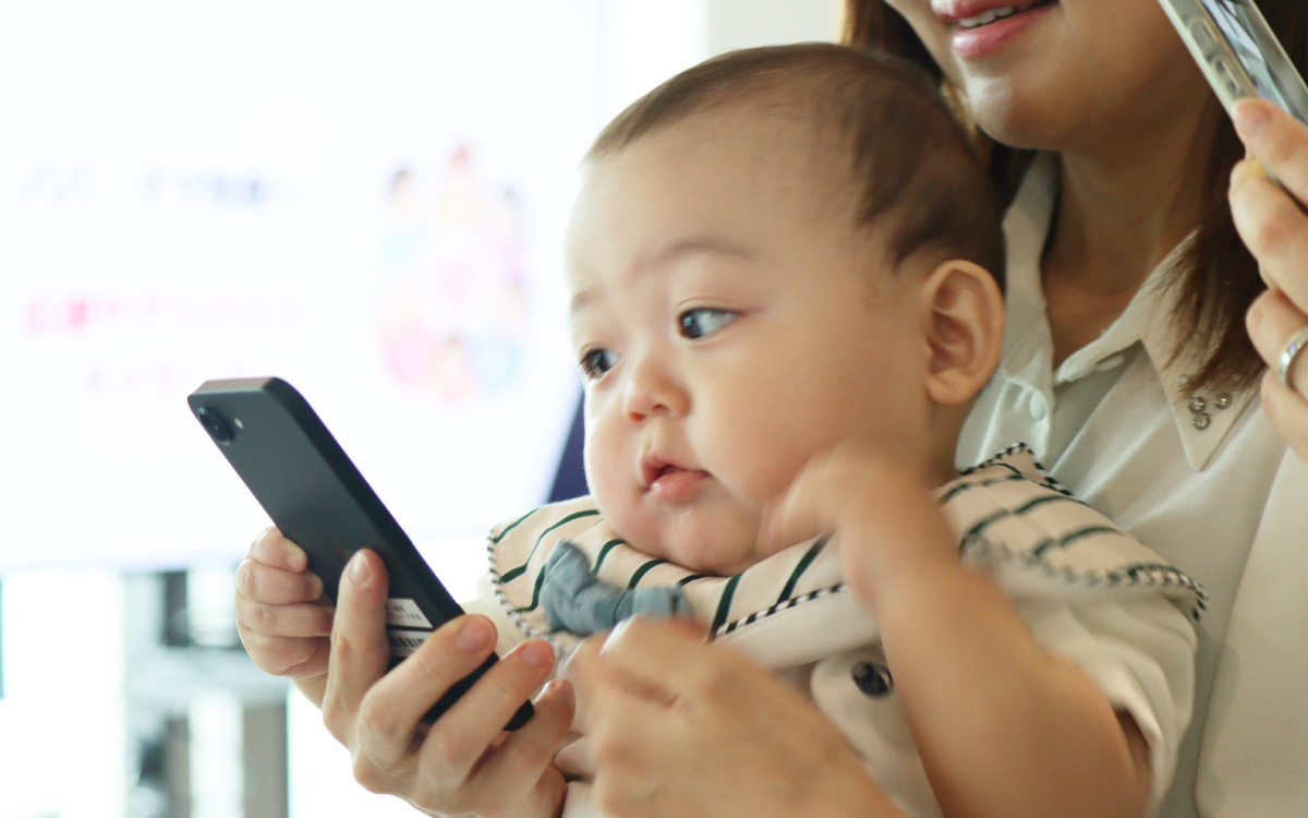 : 女性に抱っこされた赤ちゃんが、スマートフォンをじっと見つめている写真