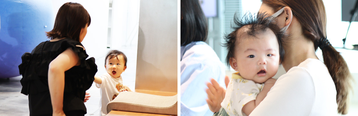 黒いノースリーブの服を着た女性が立っており、その前にいる幼い子どもが口を大きく開けて驚いたような表情を見せている様子。女性に抱っこされた赤ちゃんのバストショットである。赤ちゃんは柔らかい素材の淡い色の服を着ており、少し立った髪が印象的。