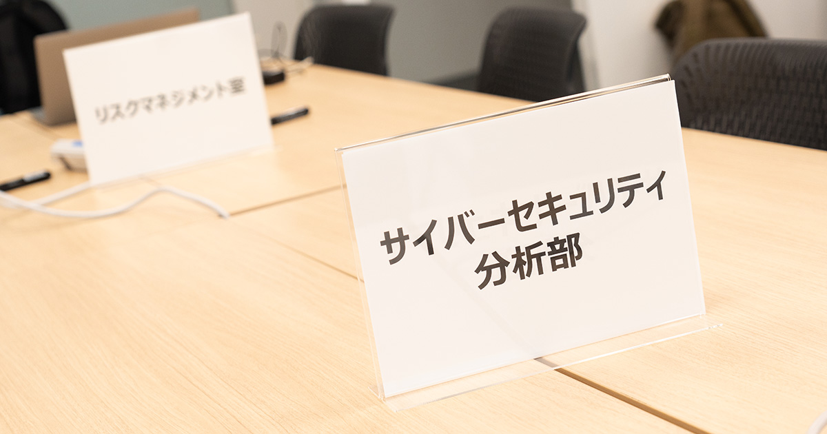 テーブル上に「サイバーセキュリティ分析部」と「リスクマネジメント室」のプレートが置かれています。