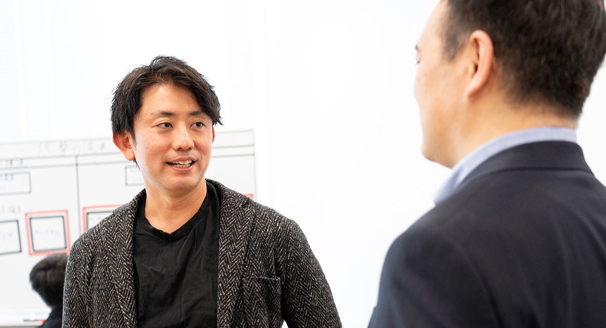 ジャケットを着た男性が、もう一人の男性と向かい合い、話をしています。背景にはホワイトボードがあります。